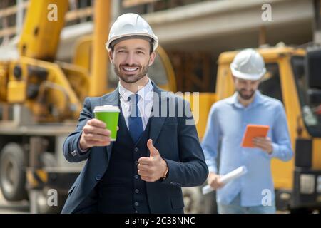 Cantiere. Supervisore della costruzione che tiene la tazza del caffè, mostrando il pollice in su, caposquadra in piedi dietro di lui che controlla qualcosa sul tablet Foto Stock