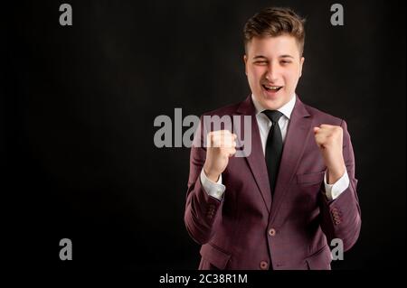 Studente di legge con capelli biondi vestiti in giacca borgogna, camicia bianca e cravatta nera si è alzato pugni esclamando con gioia ed eccitazione che si pongono su isolato Foto Stock