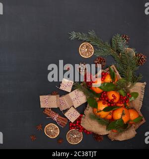 Ferme: Bacche, mandarini con foglie su vassoio di legno con tovagliolo ruvido da tela, scatole regalo, spezie e rami di abete su sfondo scuro Foto Stock