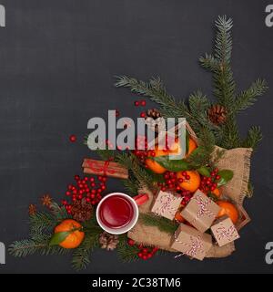 Ferme: Bacche, mandarini con foglie su vassoio di legno, tè alla frutta su tazza rossa, scatole regalo, spezie e rami di abete su sfondo scuro. V. Superiore Foto Stock