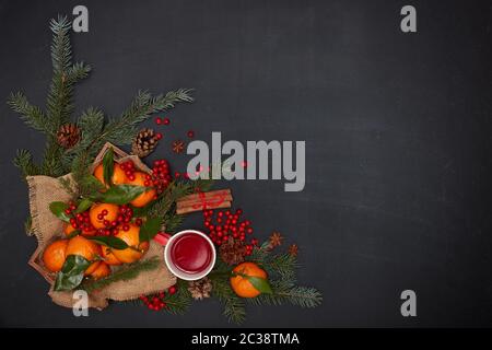 Ferme: Bacche, mandarini con foglie su vassoio di legno, tè alla frutta su tazza rossa, spezie e rami di abete su sfondo scuro. Vista dall'alto con COP Foto Stock