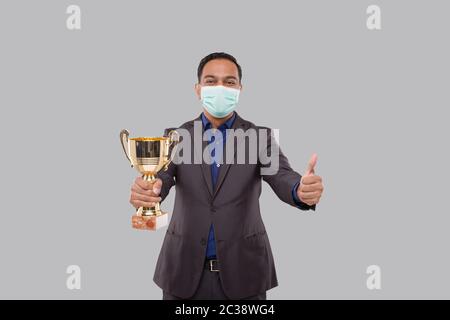 Businessman Holding Trophy indossare maschera medica mostrando Thumbp Up. Uomo d'affari indiano in piedi con Trofeo in mani Foto Stock