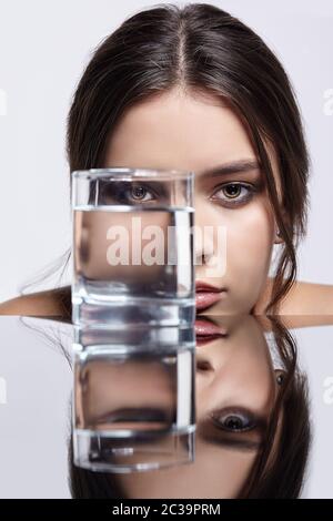 La ragazza nasconde il suo volto dietro un bicchiere d'acqua. Ritratto di bellezza della giovane donna al tavolo a specchio. Foto Stock