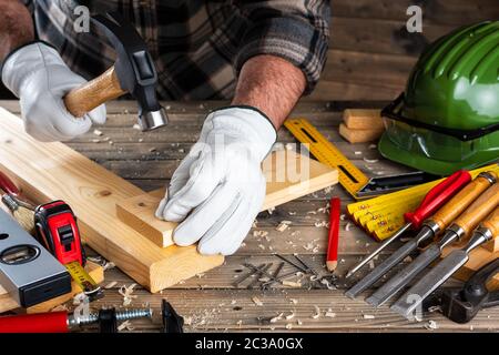 Close-up. Carpenter con le mani protette da guanti, con martello e chiodi fissa una tavola di legno. Industria edile, fai da te. Lavori in legno Foto Stock