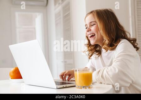 Foto di ragazza simpatica allegra che scrive sul computer portatile e ride mentre beve succo in cucina bianca Foto Stock