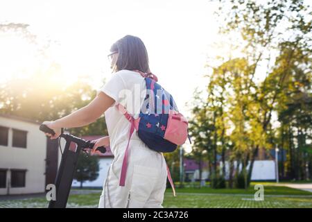 Bella giovane studentessa in occhiali che cavalcano scooters elettrico blu nel parco Foto Stock
