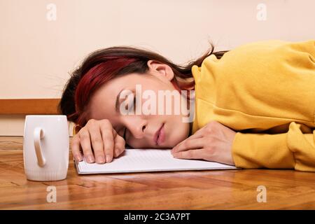 Giovane donna dormire su un notebook aperto sul pavimento con una tazza di caffè accanto a lei. Giovane e bella bruna ragazza degli studenti si è addormentato sul pavimento Foto Stock