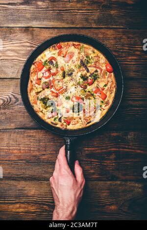 Frittata di verdure con broccoli, il peperone rosso e erbe aromatiche. Mano trattiene padella in ghisa Foto Stock