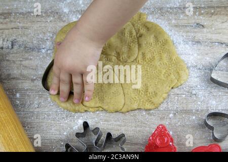 Un bambino ha messo a mano i biscotti di zenzero dall'impasto laminato, taglierina forma un abete su un fondo di legno scuro. Assistente di piccola mamma. Natale appartamento posa. Foto Stock