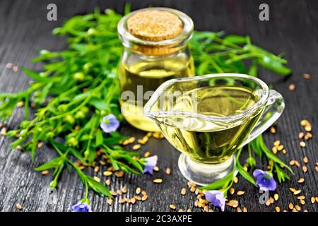 Olio di semi di lino in un vasetto di vetro e salsiera con semi, foglie e fiori di lino su una tavola di legno sfondo Foto Stock
