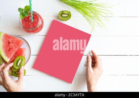 Bockup sul tema del cibo sano. Frullato di anguria, fette di kiwi e cocomero sul tavolo. Ragazza tiene un libro nelle sue mani Foto Stock