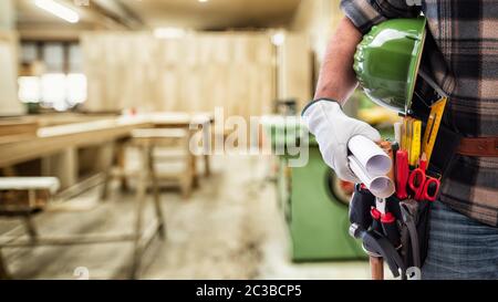 Close-up. Carpenter con le mani protette da guanti trattiene il casco e il progetto. Industria edile, laboratorio di falegnameria. Foto Stock