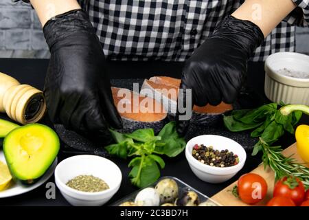 Mani preparare la bistecca di salmone per la cottura, accanto alla tabella di basilico, rosmarino spezie e peperoni, pomodori e avocado e uova di quaglia tutti steso su un b Foto Stock