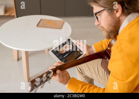 Giovane uomo serio in jumper giallo che si piega sulla sua chitarra mentre usa il touchpad per cercare le regole di sintonizzazione strumento musicale Foto Stock