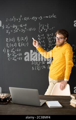 Insegnante fiducioso con pezzo di gesso che punta a una delle formule sulla lavagna mentre lo spiega agli studenti durante la lezione online Foto Stock