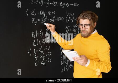 Insegnante serio con copybook che punta all'equazione sulla lavagna con pezzo di gesso mentre spiega gli studenti in linea come risolverlo Foto Stock