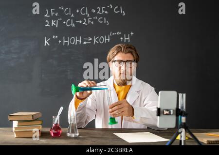 Insegnante di chimica che mostra la reazione chimica durante la miscelazione di due sostanze liquide durante il lavoro di laboratorio online davanti alla fotocamera dello smartphone Foto Stock