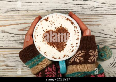 Vicino a due a due mani donna attesa e abbraccio pieno grande tazza di cappuccino cappuccino con caffè al cioccolato a forma di cuore sulla schiuma di latte su bianco tavolo in legno, eleva Foto Stock