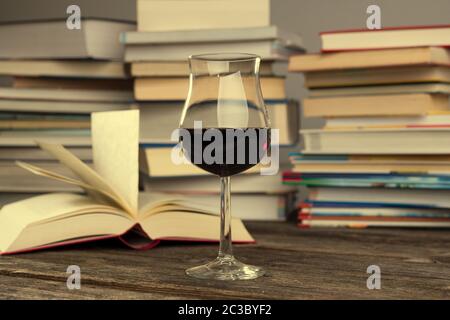 libro e bicchiere di vino davanti a mucchi di libri diversi su tavolo di legno Foto Stock