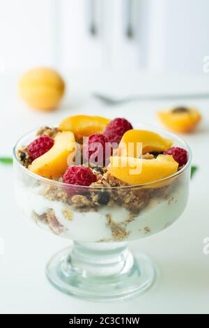 Parfait sano con yogurt greco, muesli fatti in casa, fette di albicocca e lamponi in un calice di vetro. Sfondo grigio di legno con albicocche Foto Stock