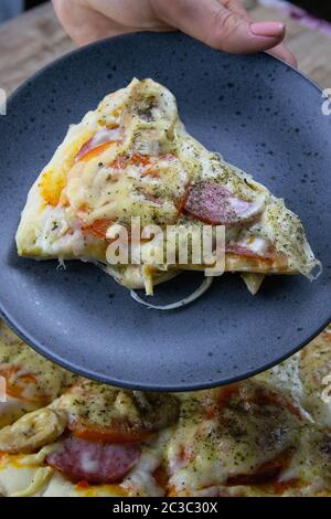 La mano trattiene una piastra con un pezzo di pizza oltre la pizza che sorge su una tavola di legno. Pizza con mozzarella di bufala, salame, pomodori e banane Foto Stock