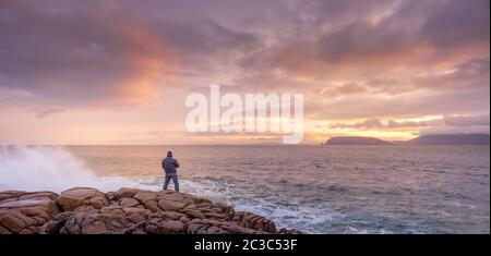 Fotografo che fotografa le onde che si infrangono sulle rocce con un'incredibile alba sullo sfondo. Foto Stock