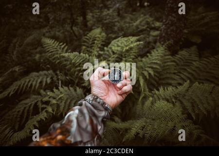 Bussola in una mano con sfondo di foglie di felce sfocate. Escursionista alla ricerca di direzione nella foresta selvaggia. Spazio di copia. Foto Stock