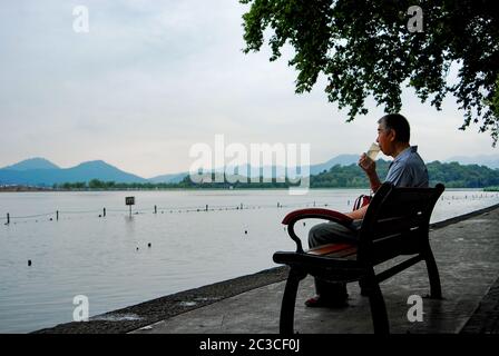 Un uomo anziano seduto alla panchina beve il tè da una bottiglia di plastica riutilizzabile mentre si guarda il Xihu, le acque del Lago Occidentale in giornata nuvolosa Foto Stock