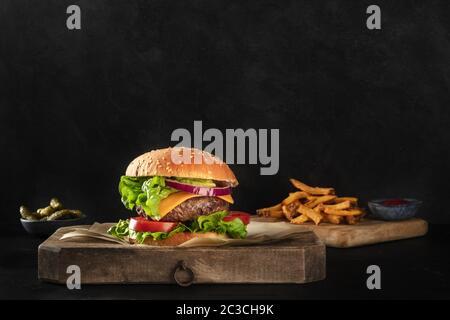 Burger con manzo, formaggio, cipolla, pomodoro e insalata verde, vista laterale su sfondo scuro con patatine fritte e sottaceti, con un Foto Stock