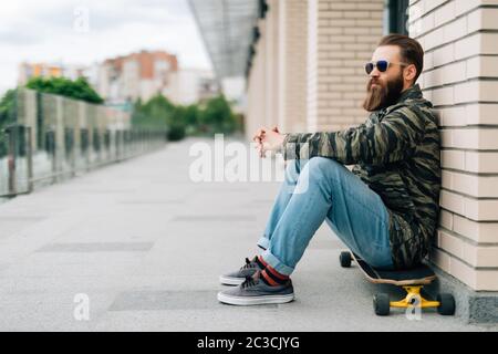 Bel giovane seduto sul longboard sulla strada in città. Concetto di skateboard urbano. Foto Stock