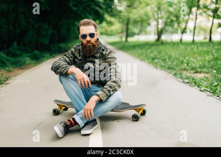 Giovane uomo con palchi in abiti casual seduto sulla tavola lunga o skateboard all'aperto. Concetto urbano, sottocultura, skateboard Foto Stock