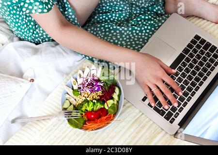 Donna rilassata che usa il computer portatile mentre si siede a letto e si ha un sano pranzo vegetariano a casa Foto Stock