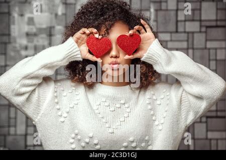 Una giovane ragazza afro-americana con capelli ricci in un maglione bianco tiene due cuori nelle sue mani . chiude gli occhi con i cuori e fa Foto Stock