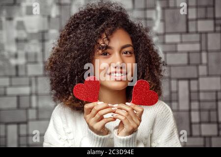 Ritratto in primo piano di una giovane ragazza afro-americana con capelli ricci in un maglione bianco che tiene due cuori di carta . profondità poco profonda di Foto Stock