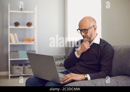 Uomo anziano con il laptop che comunica online, lavorando, consultando il medico o prendendo corso di formazione a casa Foto Stock