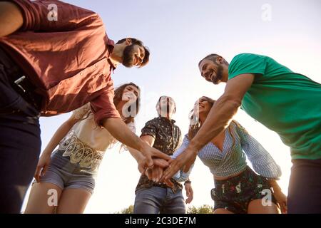 Vista ad angolo basso di giovani amici sorridenti che mettono le mani insieme in cerchio, all'aperto. Concetto di unità o di supporto Foto Stock