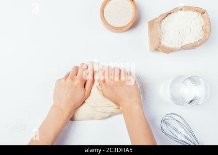 Mani femminili impastare l'impasto su un tavolo bianco accanto a ingredienti e utensili, vista dall'alto. Foto Stock