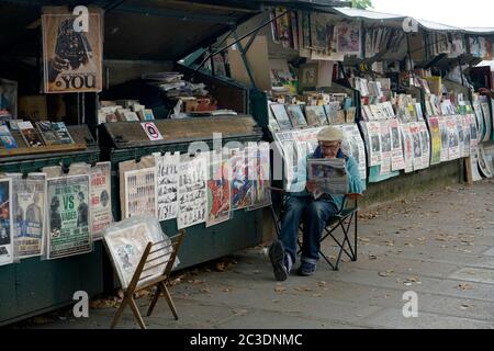 Un maschio Bouquiniste -il giornale di lettura del bookseller antico davanti alle sue scatole di esposizione lungo la riva della Senna River.Paris.France Foto Stock