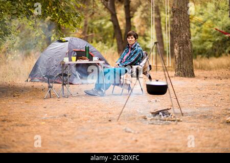 Donna anziana che prepara il cibo sul fuoco mentre vive nella vita della tenda nella foresta. Foto Stock