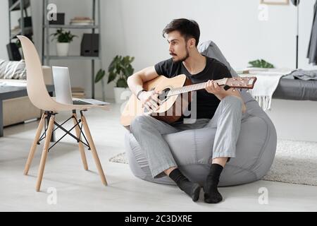 Uomo giovane e bearded concentrato seduto in borsa e guardare video sul portatile mentre si impara a suonare la chitarra Foto Stock