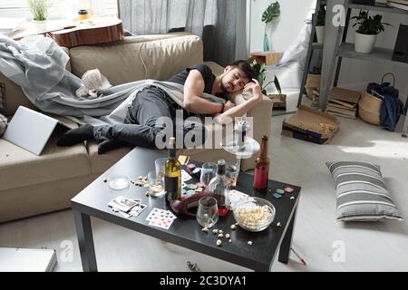Un giovane ubriaco che dorme sul divano tra le cose dissuasche nella camera sporca con una bottiglia di alcool sul tavolo Foto Stock