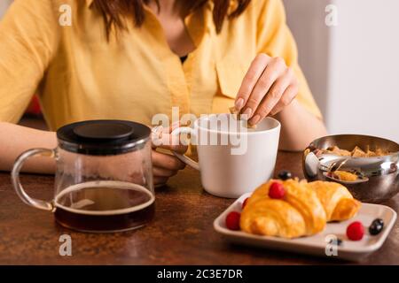 Mani di giovane donna che mettono due cubetti di zucchero di canna in tazza con tè o caffè mentre andando fare colazione la mattina Foto Stock