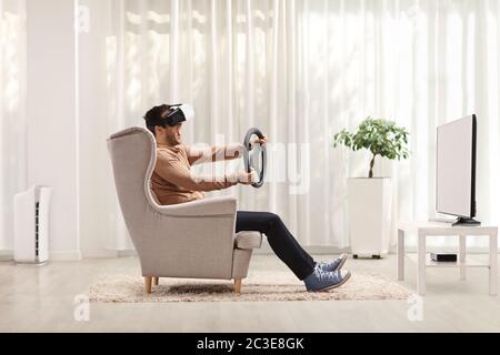 Un giovane seduto in poltrona utilizzando una cuffia per realtà virtuale e tenendo il volante a casa Foto Stock