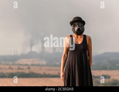 Giovane ragazza in un vestito nero e maschera a gas sullo sfondo di fumo fabbrica camini in Ucraina primo piano Foto Stock