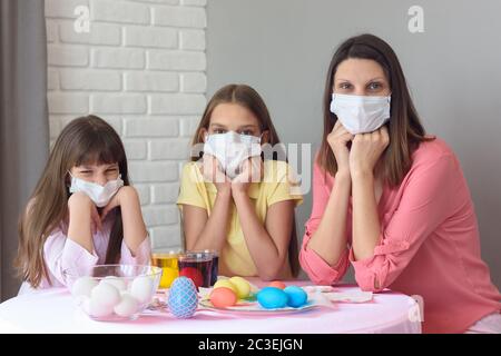 Mamma e due figlie al tavolo in maschere mediche dipingono le uova di Pasqua per la vacanza Foto Stock