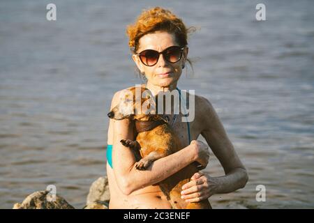 Donna caucasica matura si siede in acqua sulla pietra e tenere il cane di razza Dachshund in armi. Caldo a tema e vacanza estiva. Vacanze in spiaggia sul fiume con Foto Stock