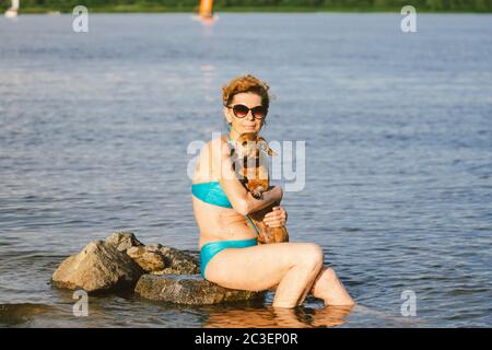 Donna caucasica matura si siede in acqua sulla pietra e tenere il cane di razza Dachshund in armi. Caldo a tema e vacanza estiva. Vacanze in spiaggia sul fiume con Foto Stock