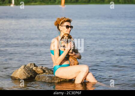 Donna caucasica matura si siede in acqua sulla pietra e tenere il cane di razza Dachshund in armi. Caldo a tema e vacanza estiva. Vacanze in spiaggia sul fiume con Foto Stock