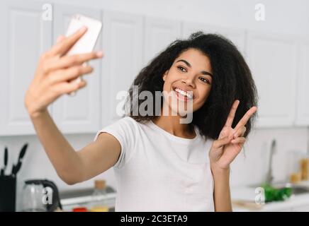 Felice bella donna afroamericana prendendo selfie sulla macchina fotografica del telefono mobile, mostrando il segno di vittoria. Video in streaming online con influencer emotivo Foto Stock