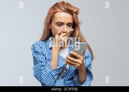 Sorpresa giovane donna con capelli biondi fragola che copre la bocca con la mano e guardando il suo telefono, riceve sms, notizie, messaggio, isolato su bac grigio Foto Stock